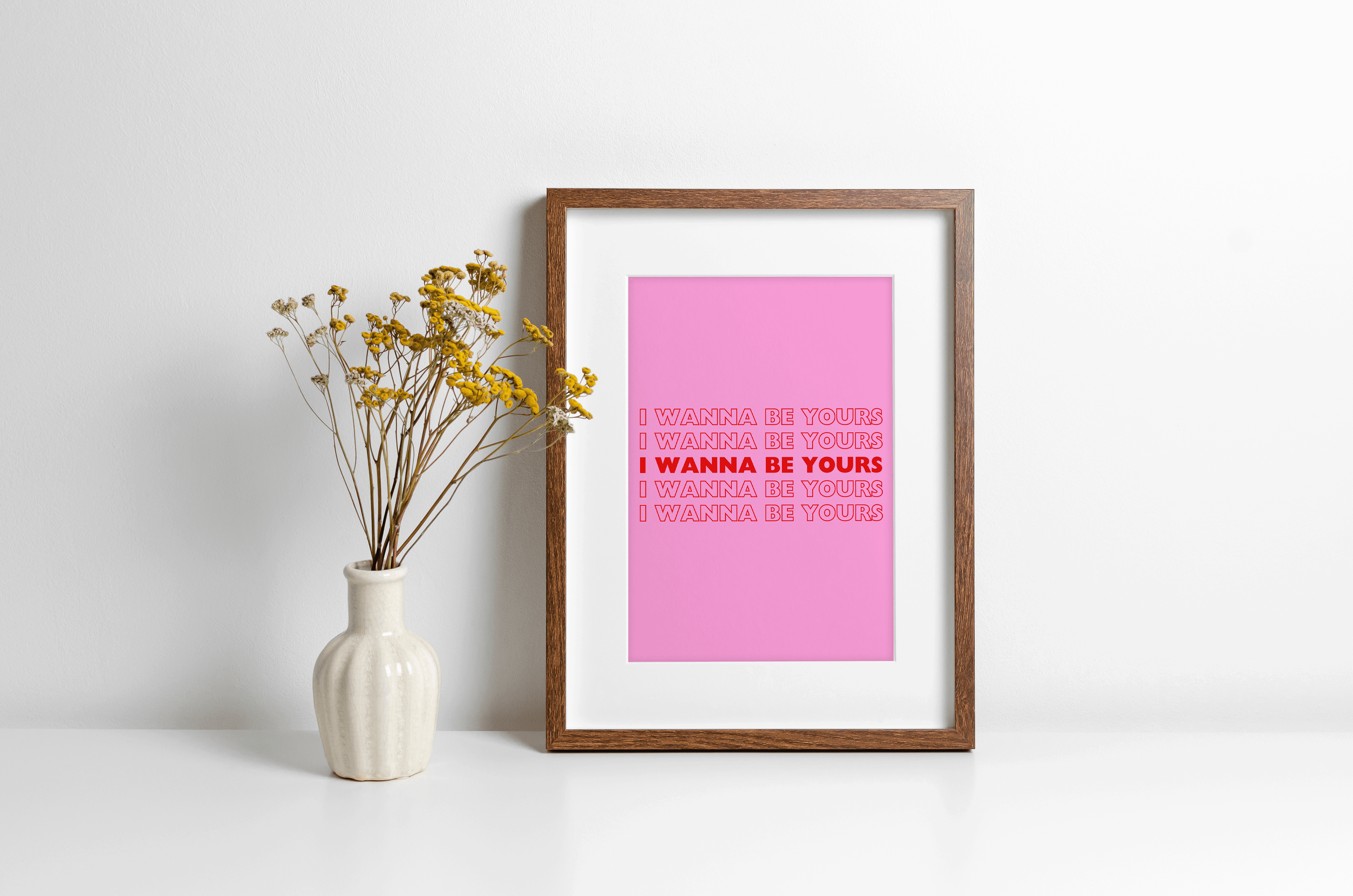 A bold music lyrics print featuring Arctic Monkeys "I wanna be yours" bold typography in red against a bright pink background. Displayed in a wooden frame against a white wall with yellow flowers in vase alongside.