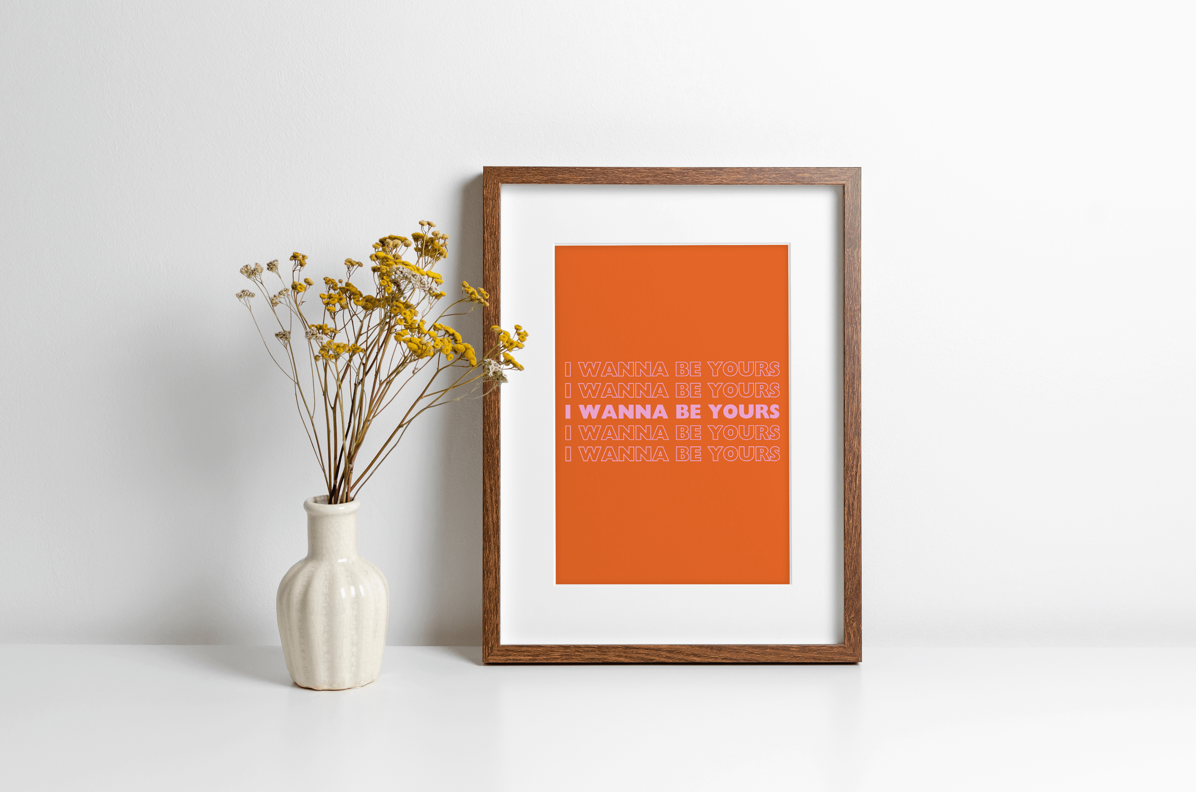 A music lyrics print featuring Arctic Monkeys "I wanna be yours" bold typography in pink against a orange background. Displayed in a wooden frame against a white wall with yellow flowers in vase alongside.