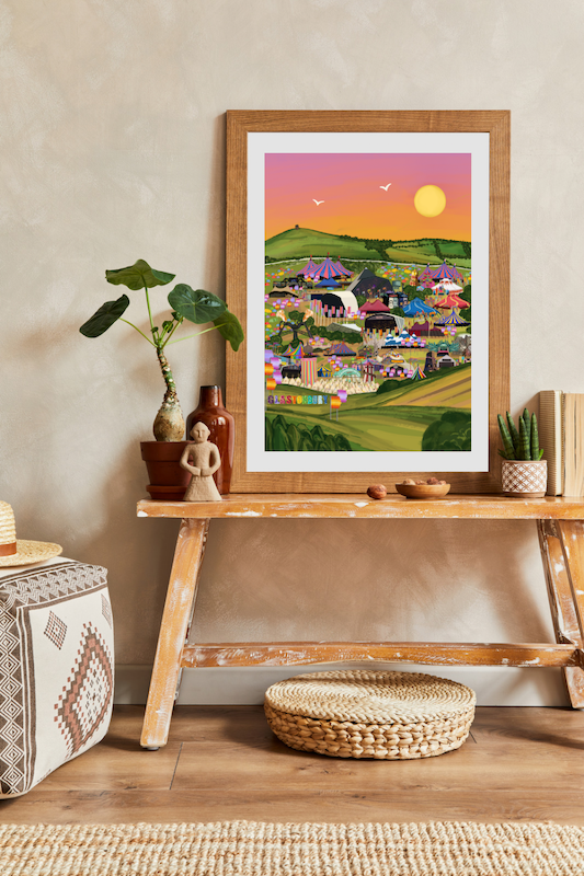 A framed Glastonbury festival print on a wooden bench, surrounded by boho decorative items and plants