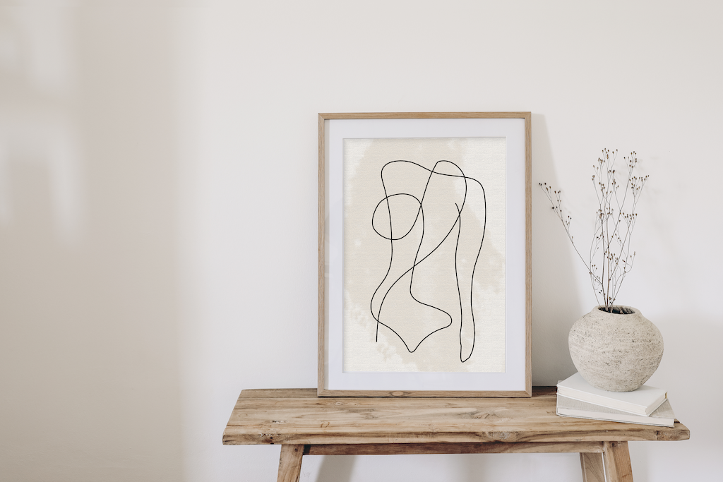 A framed print featuring a line drawing of the female body, stood on a wooden bench against a while wall alongside decorate items.