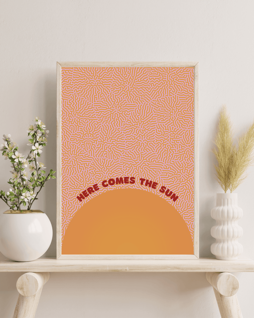 Here comes the sun art print, in orange and red colour scheme. Print is framed in a wooden frame, on a bench with indoor plants and foliage. For display purposes only.