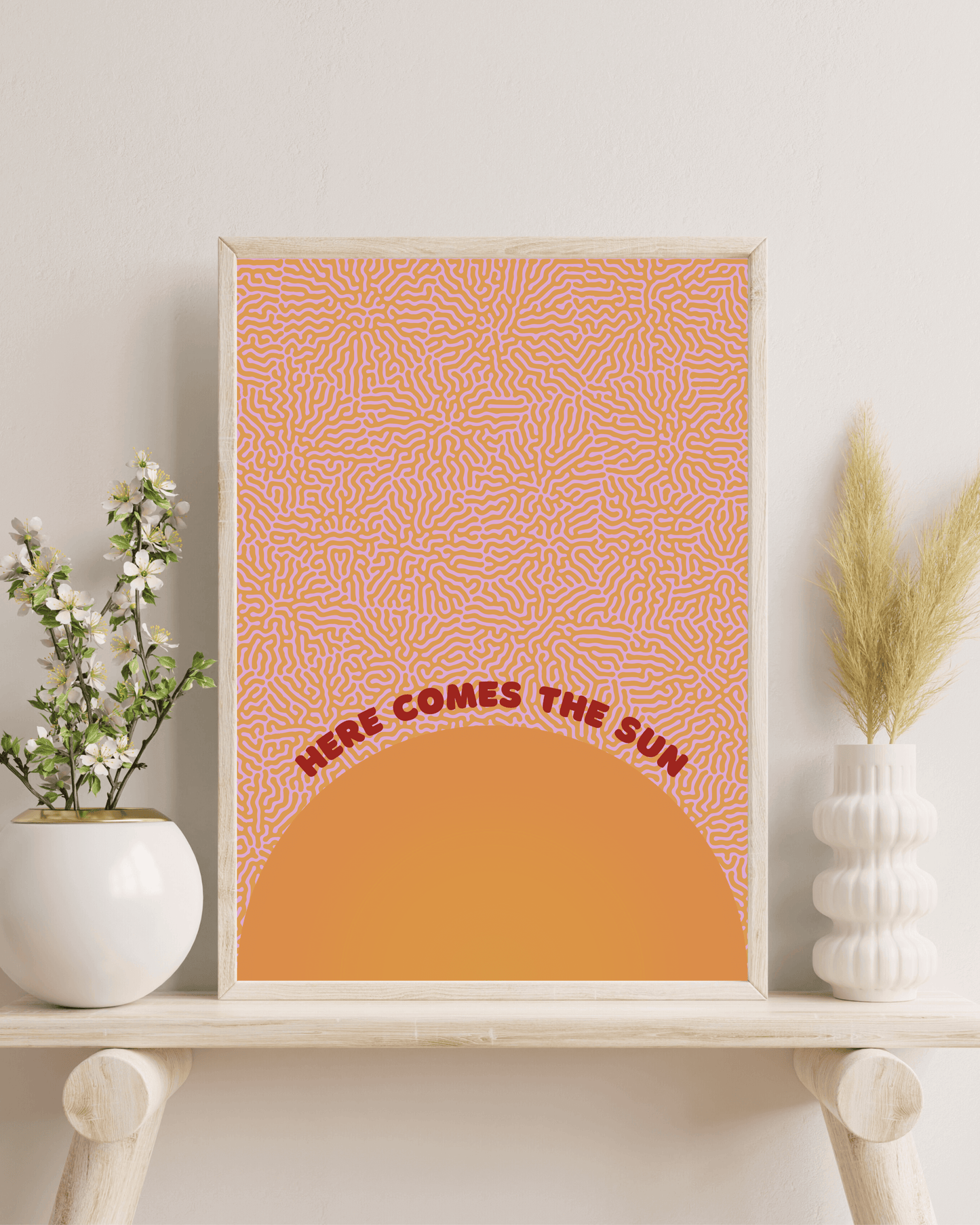 Here comes the sun art print, in orange and red colour scheme. Print is framed in a wooden frame, on a bench with indoor plants and foliage. For display purposes only.