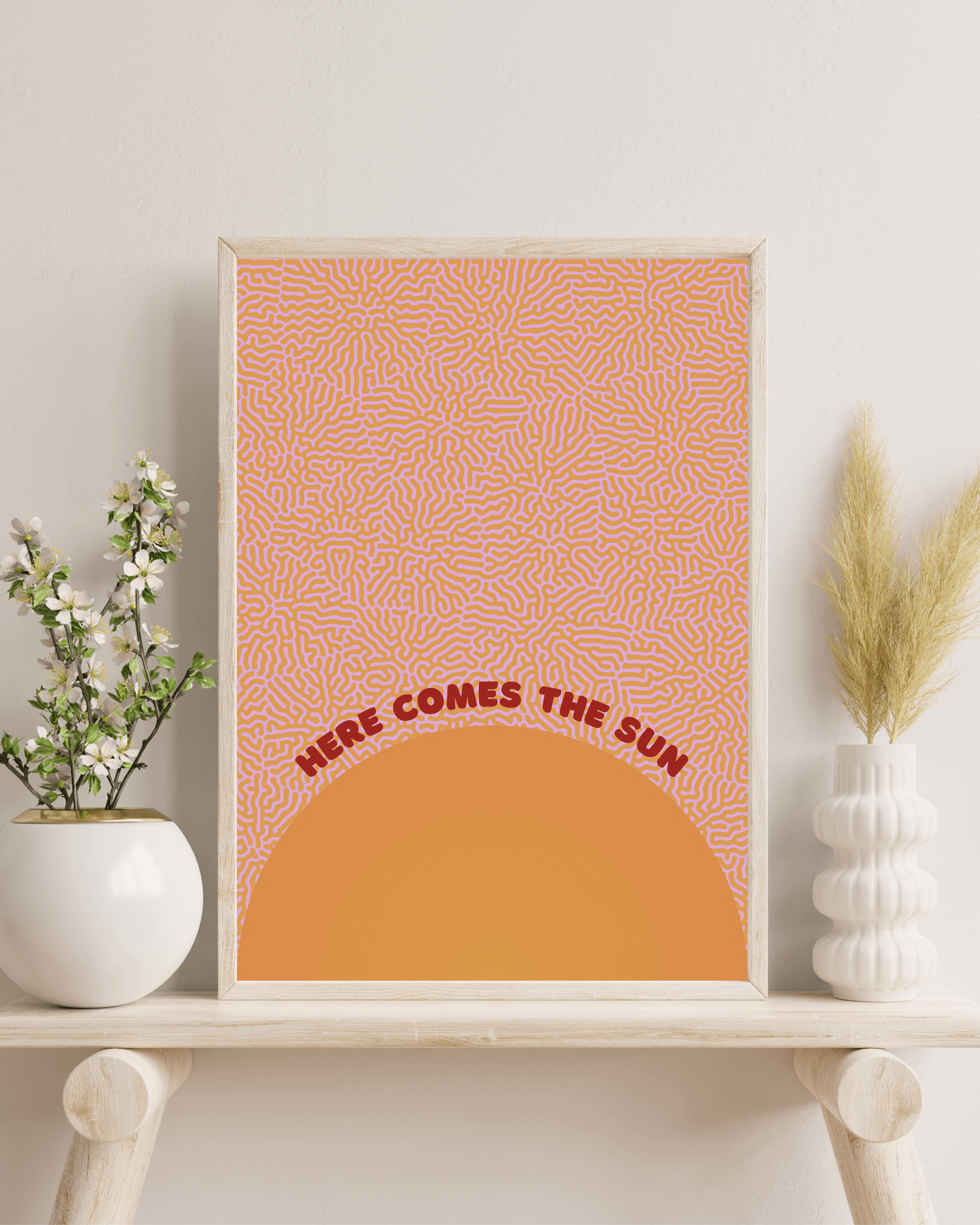 Here comes the sun art print, in orange and red colour scheme. Print is framed in a wooden frame, on a bench with indoor plants and foliage. For display purposes only.