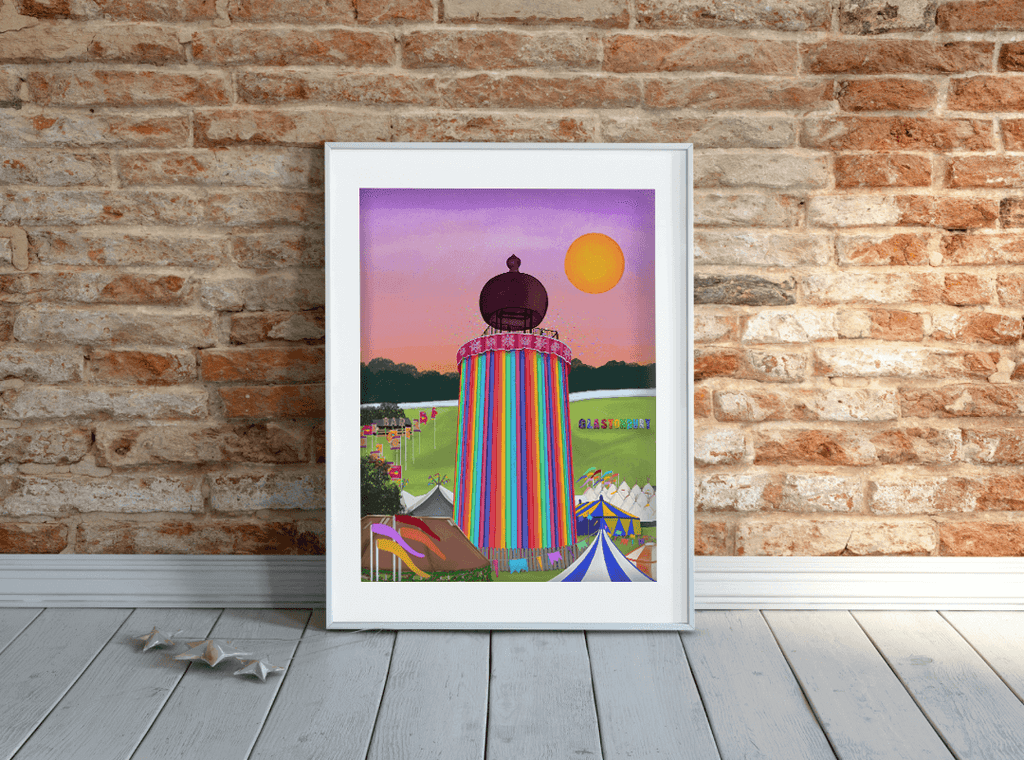 Colourful, vibrant art print of Glastonbury Festival's iconic ribbon tower. Featuring the Glastonbury sign, a large bright sun and a purple-pink sky, displayed in a white frame leaning against a brick wall.