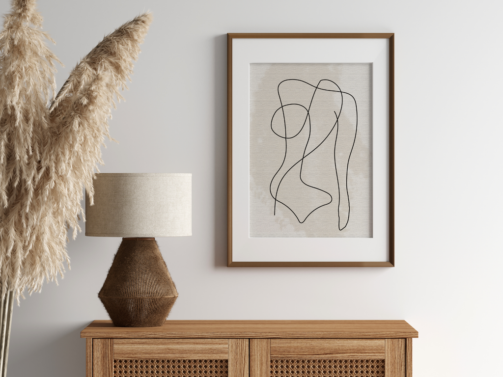 Minimalist line art print featuring a line drawing of female body, on a neutral background displayed in a wall hung wooden frame above sideboard with lamp and decorative pampas.