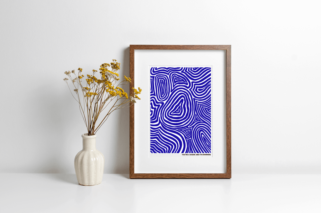 Music lyrics art print featuring a blue swirl illustration above the lyrics "you're a shark and i'm swimming" from alt-J, displayed in a wooden frame alongside a vase of yellow flowers
