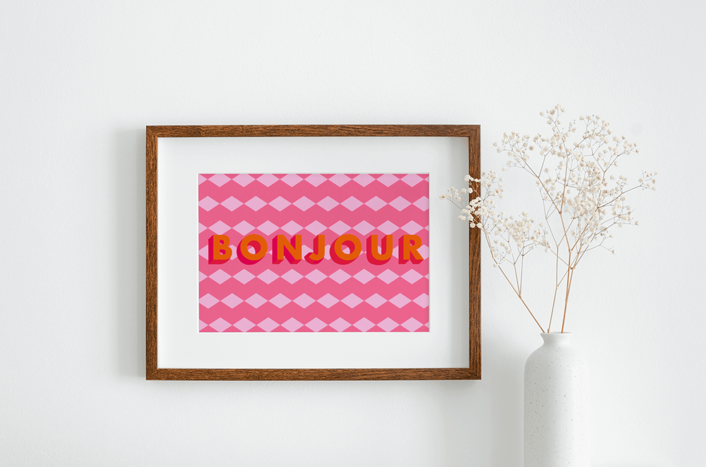 Art print with geometric pink background and orange text reading "Bonjour", hanging on a white wall next to a vase of white flowers.