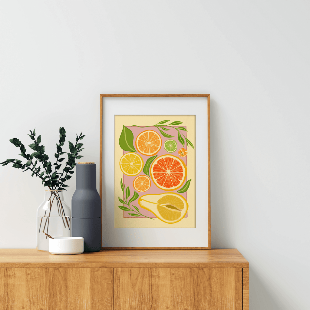 An art print featuring citrus fruits in vibrant, yellow, oranges and greens against a pink and yellow background. Displayed in a wooden frame on a cabinet surrounded by decorative items.