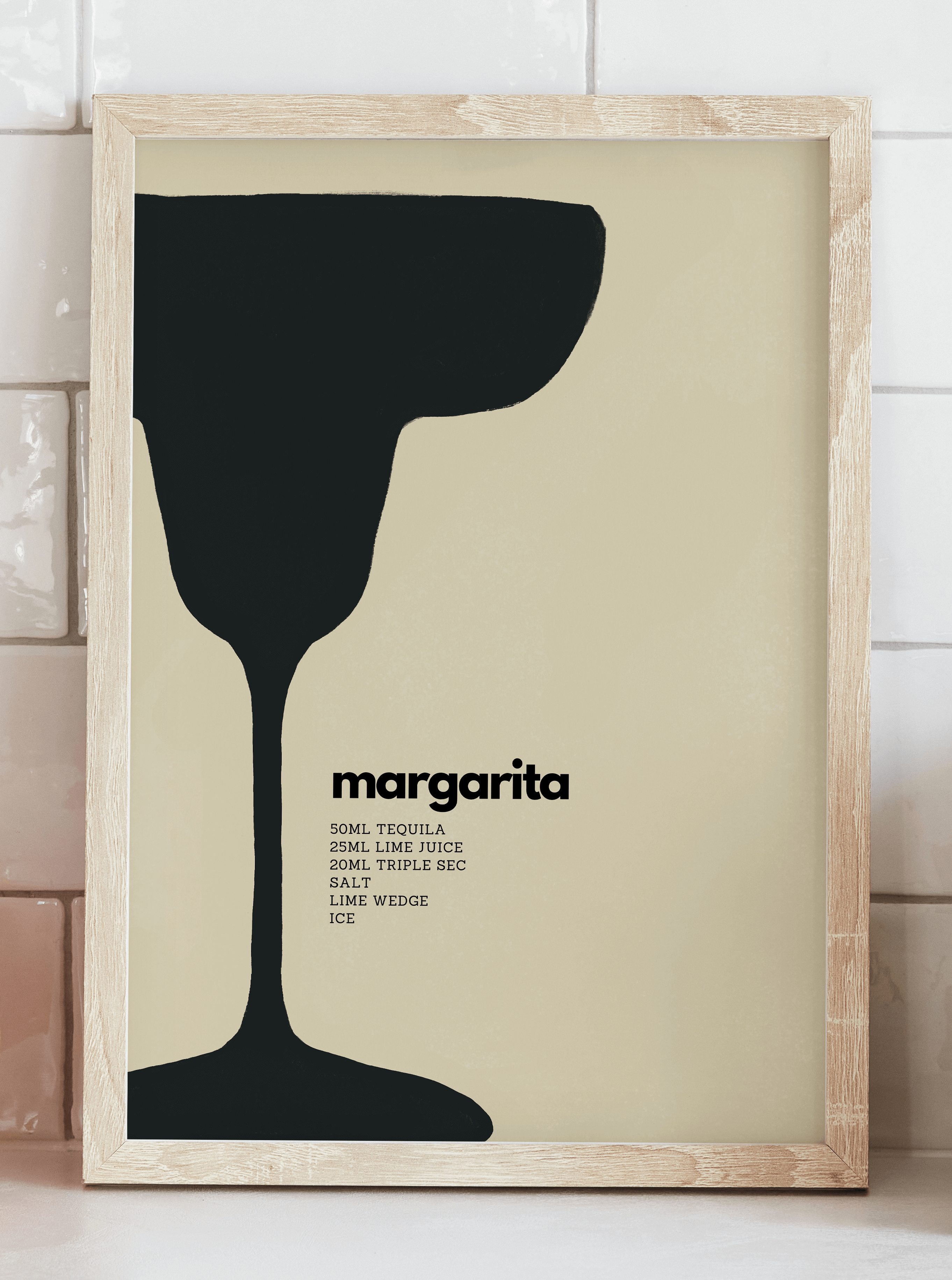 Wall art print featuring minimalist black illustration of a margarita cocktail with measurements for recipe alongside. Displayed in a pine frame leaning against a tiled kitchen wall.
