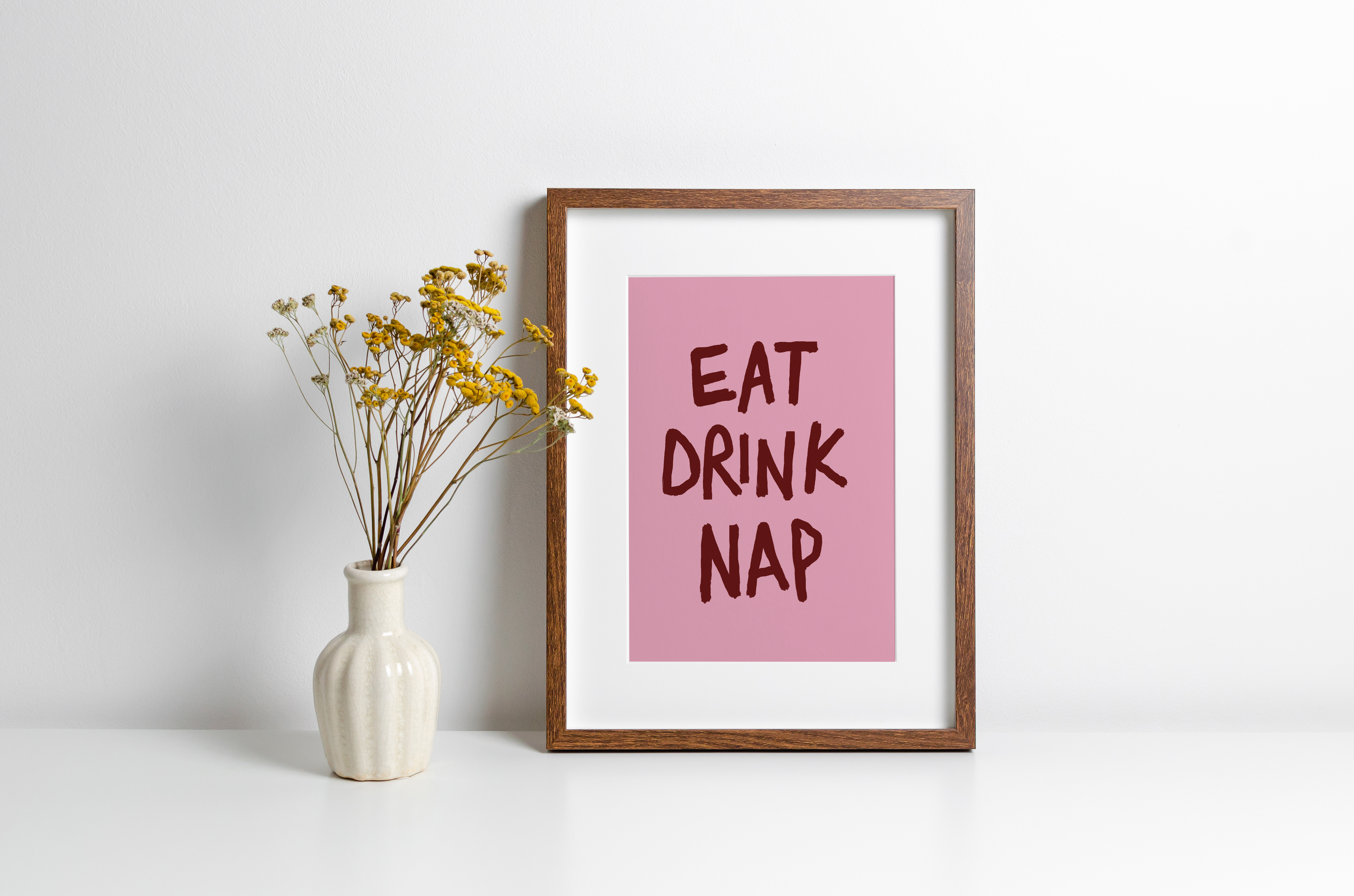Framed print with 'EAT DRINK NAP' text on a white surface with a vase of flowers.