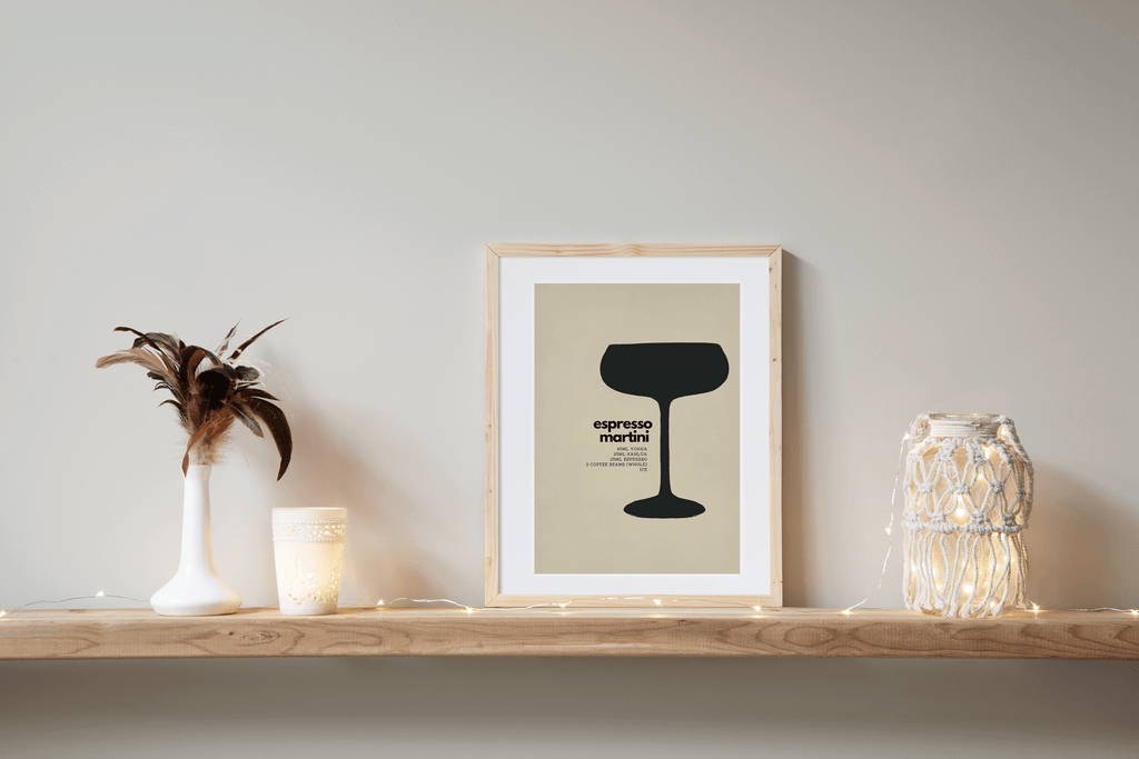 Wall art print featuring minimalist black illustration of a espresso martini cocktail with measurements for recipe alongside. Displayed in a pine frame on a shelf leaning against a wall with decorative items alongside