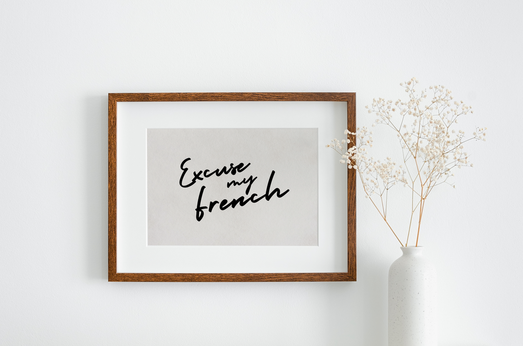 Excuse my French art print in a monochrome colour scheme. The typography is black, in a handwritten style against an off white/greige background. The print is displayed in a wall hung wooden frame, next to a white vase with foliage.