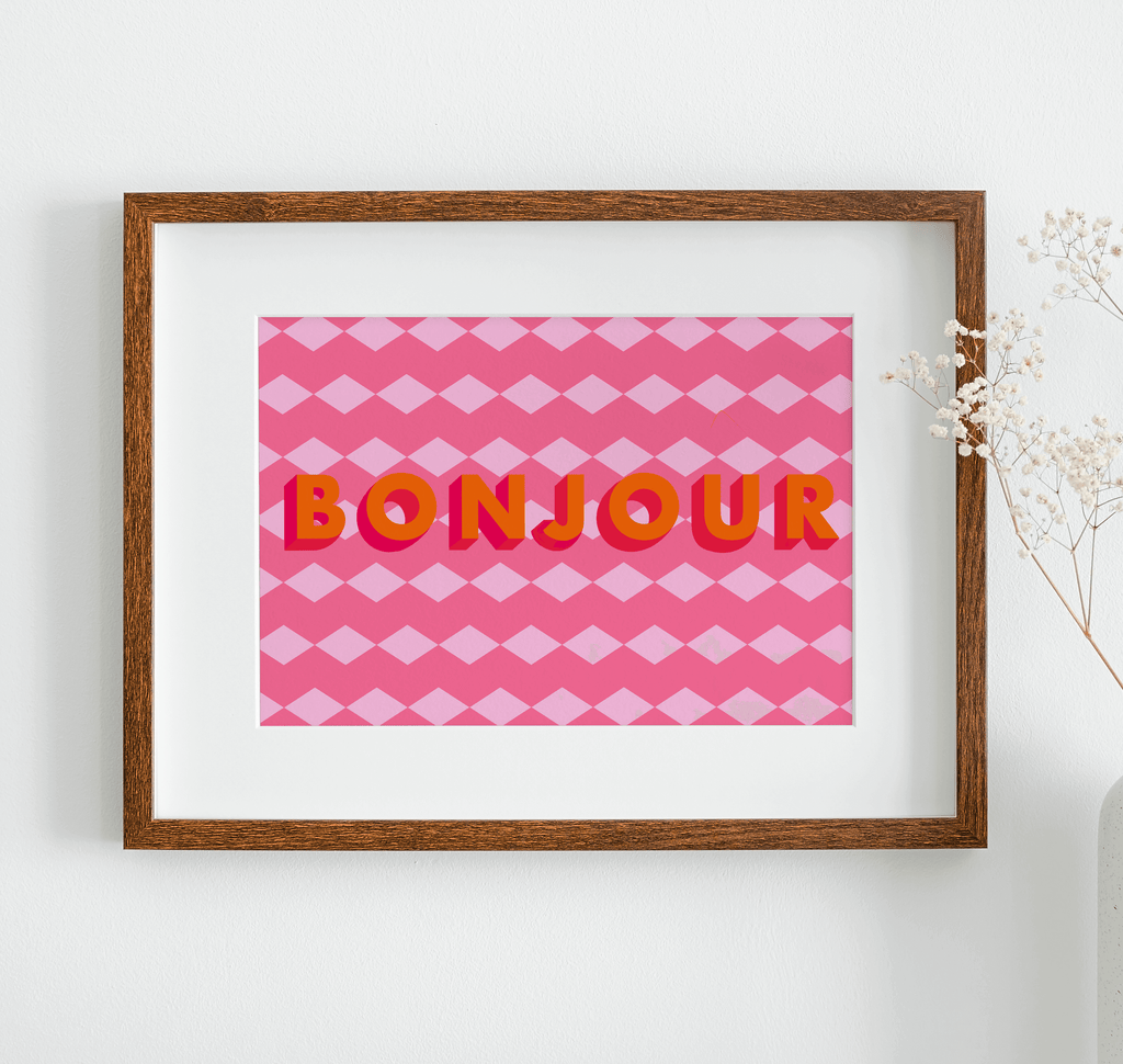 Art print with geometric pink background and orange text "Bonjour" displayed in a wooden frame hanging on a white wall