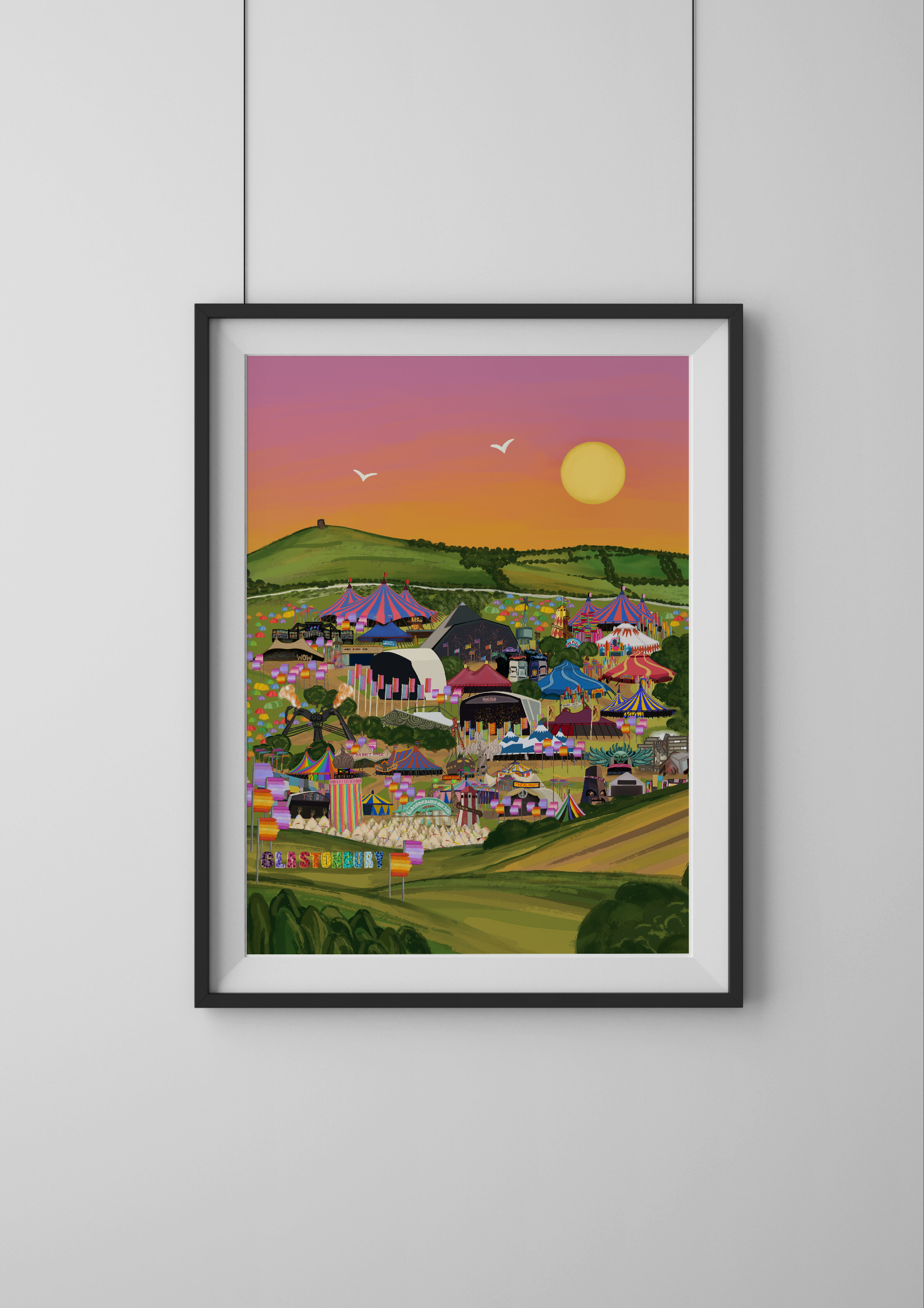 Framed artwork of a colorful festival scene with tents and greenery against a sunset sky.