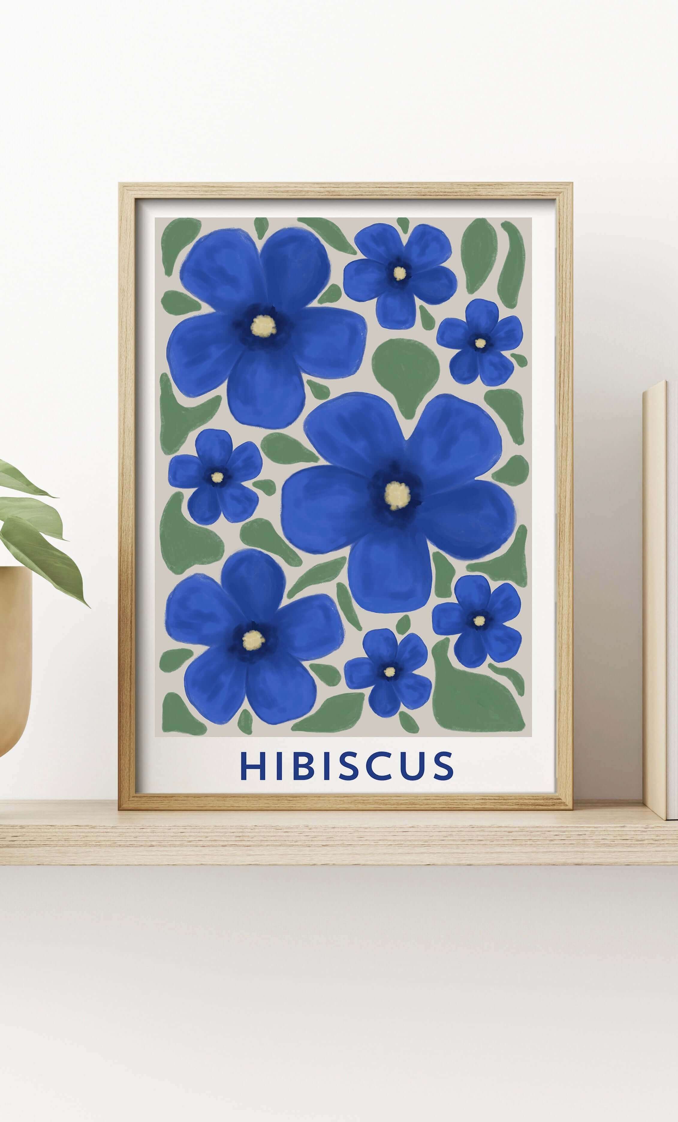 Blue hibiscus wall art print with green leaves and "hibiscus" typography, displayed in a wooden frame on a shelf alongside decorative plants.