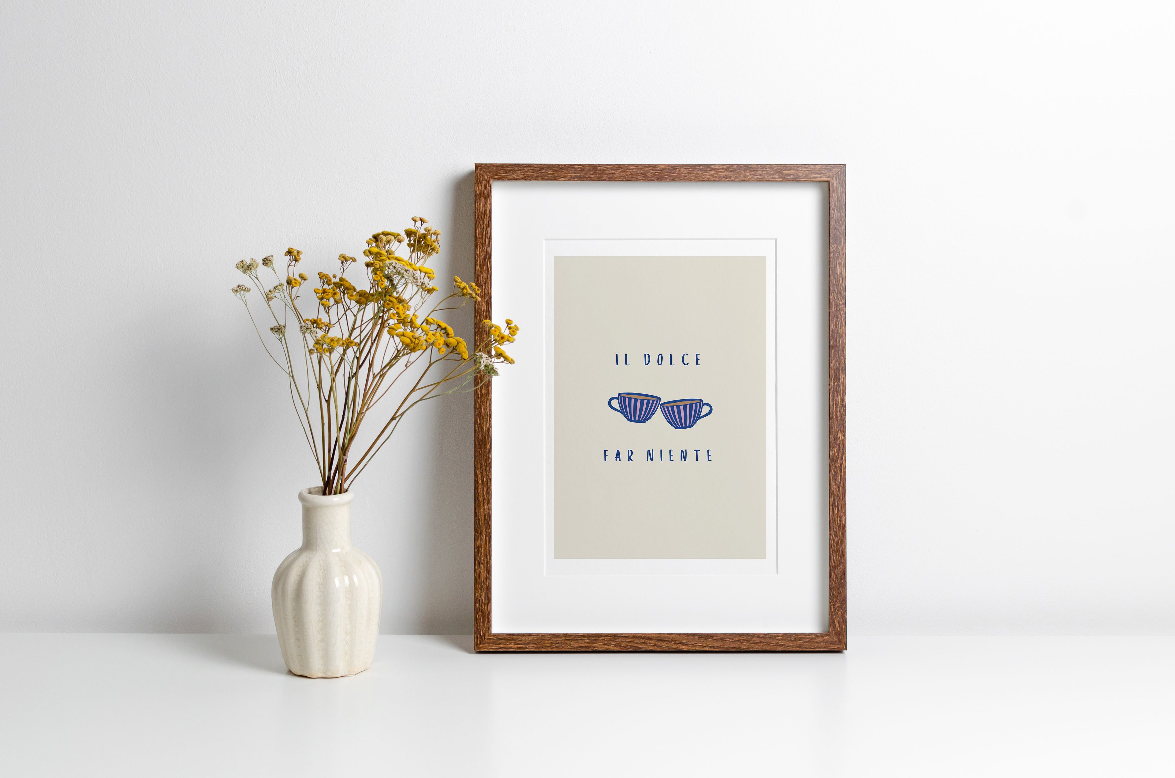 A framed kitchen print with coffee cup illustration and typography "il dolce far niente" against a beige background with white border. The print is displayed in a wooden frame against a white background alongside a vase of yellow flowers.