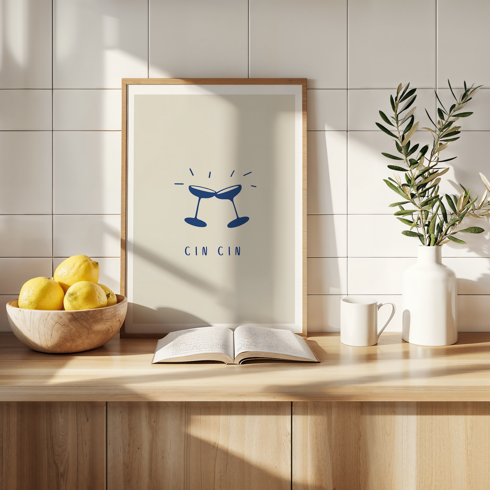 An Italian "cin cin" poster featuring two champagne or prosecco coupes clinking together against a beige background with white border. The print is displayed in a wooden frame in a kitchen surrounded by aesthetic items and fruit.