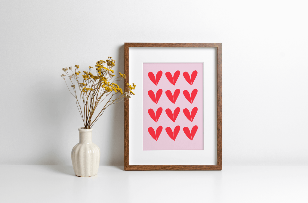 Wall art print featuring repeat heart pattern in red against a pink background, displayed in a wooden frame alongside a decorative vase with yellow flowers.