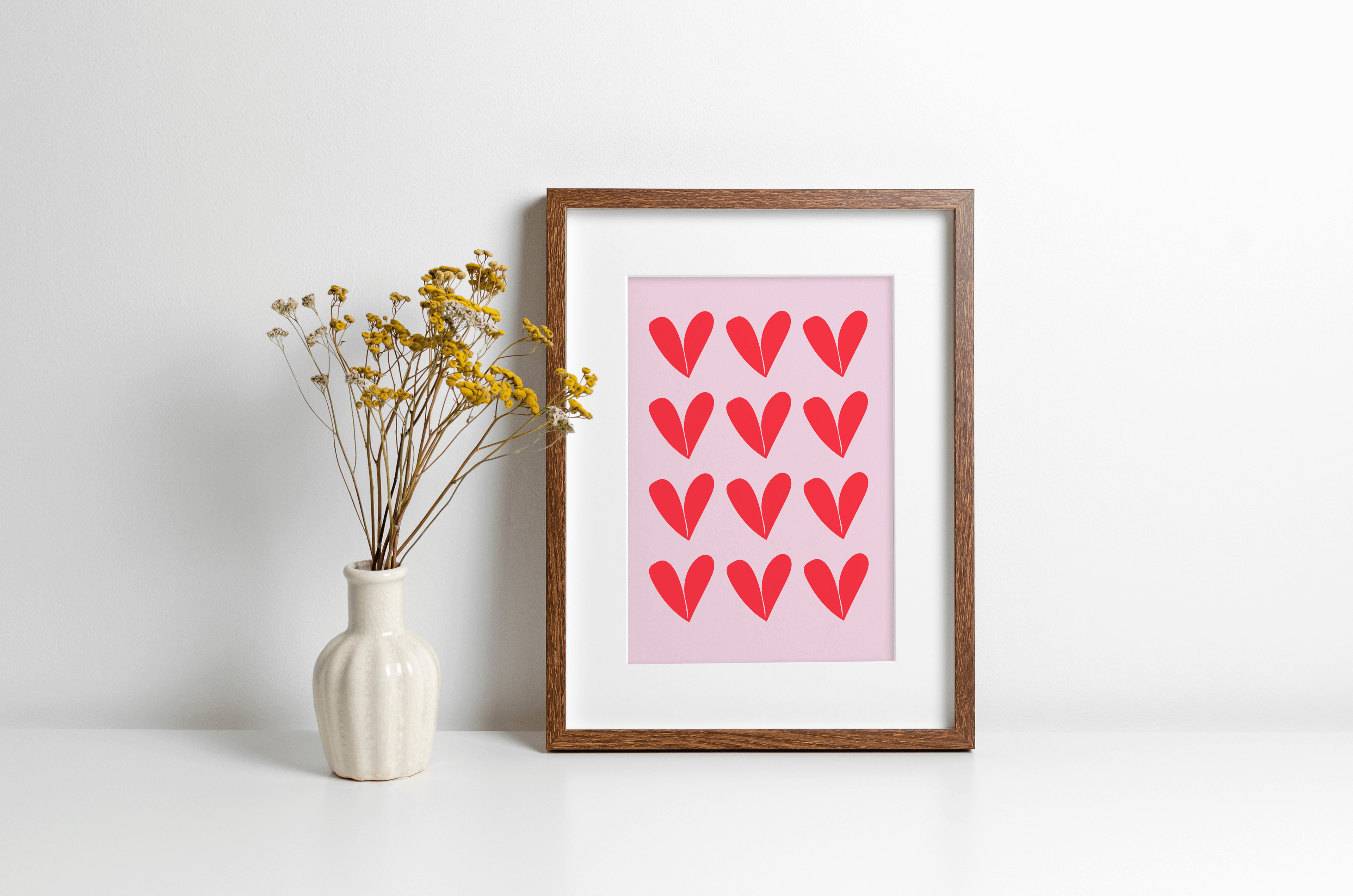 Wall art print featuring repeat heart pattern in red against a pink background, displayed in a wooden frame alongside a decorative vase with yellow flowers.