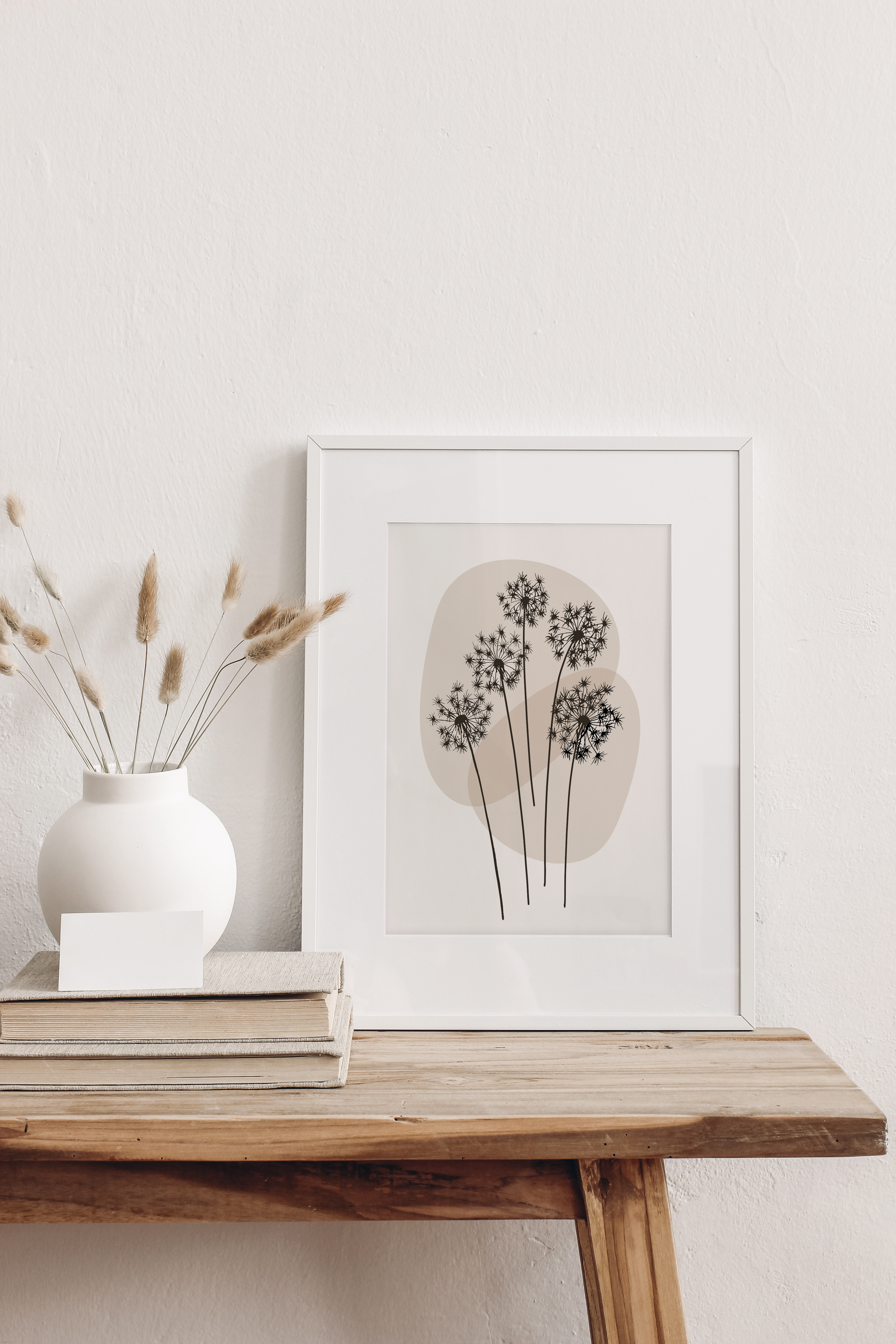 Minimalist line art print featuring a line drawing of dandelion, on a neutral background displayed in a wall leaning frame on a bench with decorative items.