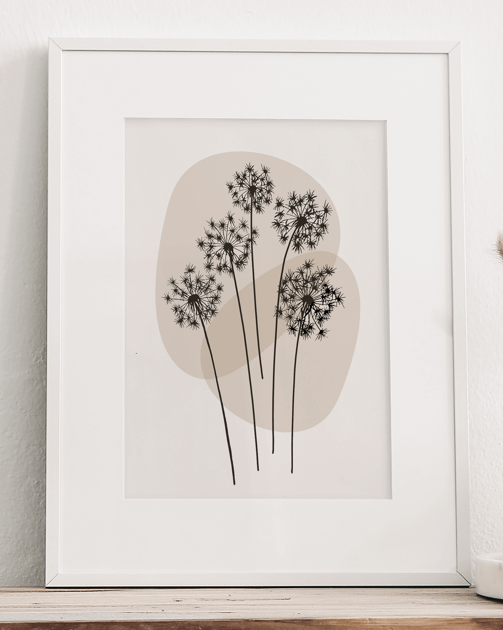 A black and white line art print of dandelions in a white frame, leaning against a wall.