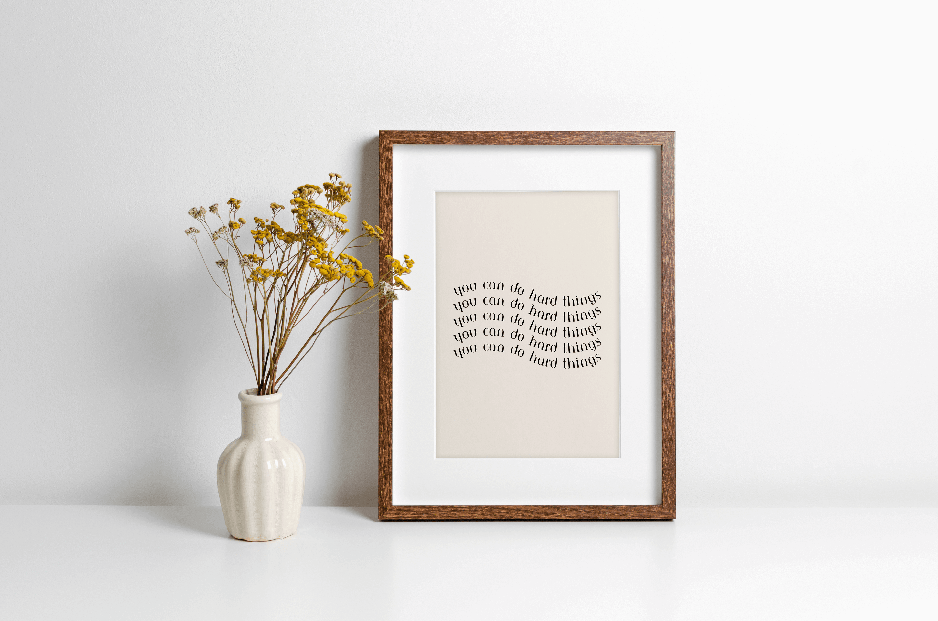 A framed typography poster with the phrase 'You can do hard things' in a monochrome, black bold typography, presented in a simple wooden frame alongside decorative vase.
