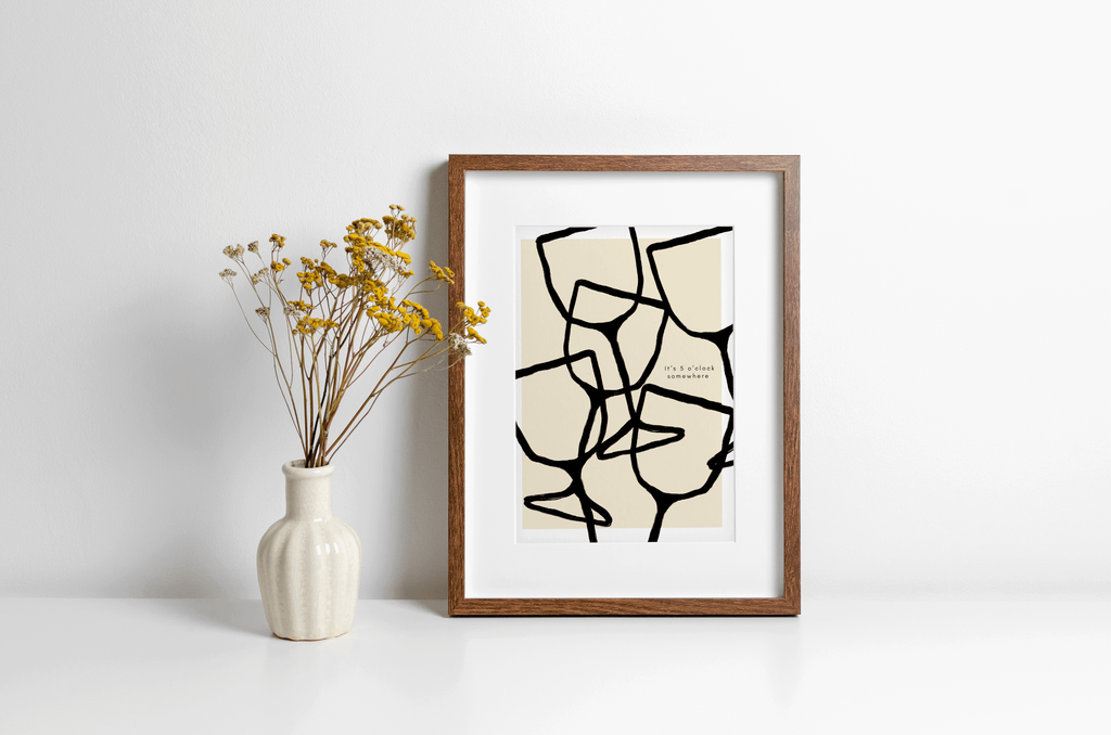 Wall art print featuring black wine pattern illustration in bold black line art and typography "it's 5 o'clock somewhere" alongside. Print is displayed in a wooden frame leaning against a wall.