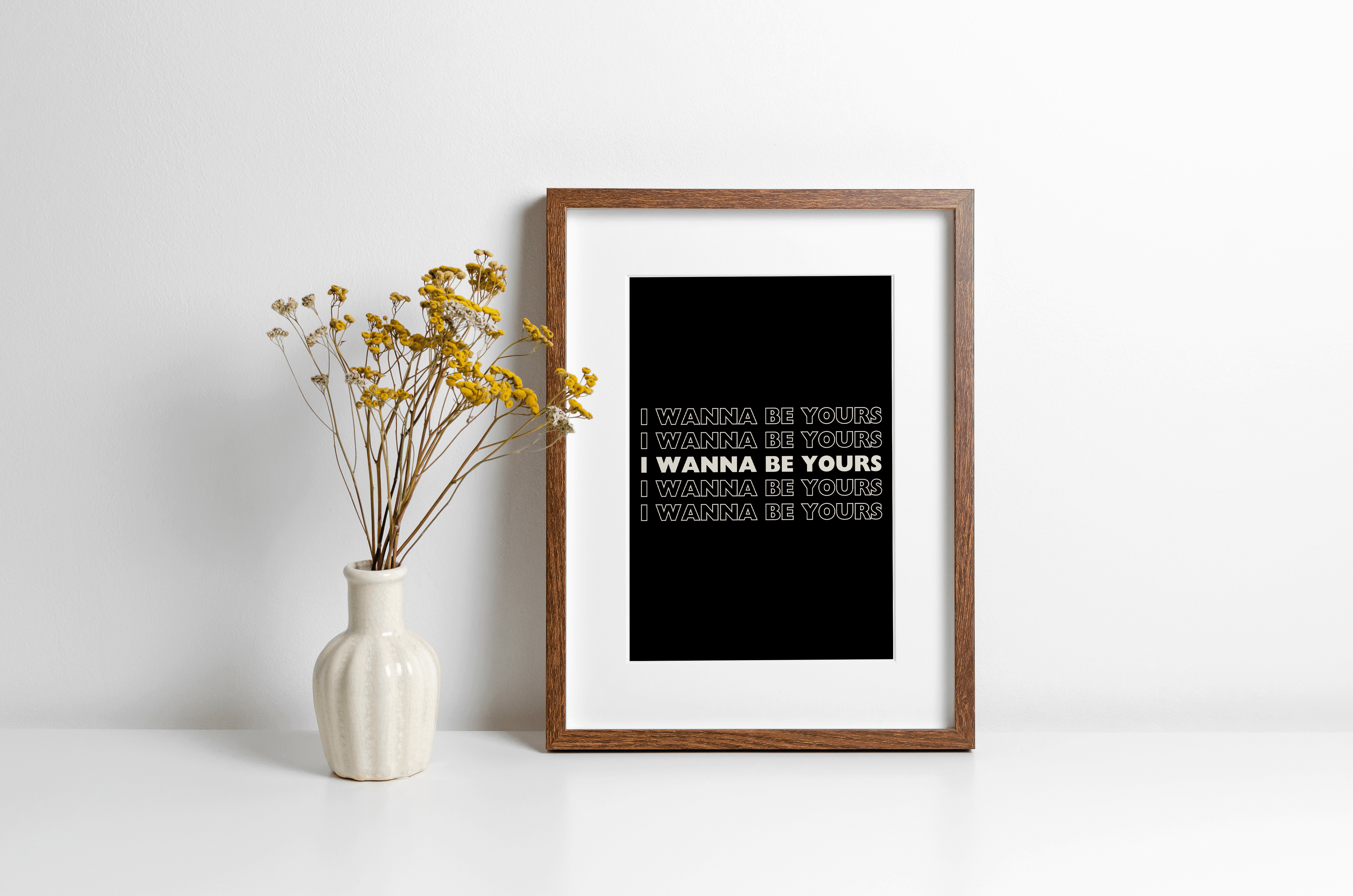 A music lyrics print featuring Arctic Monkeys "I wanna be yours" typography in white against a black background. Monochrome print is displayed in a wooden frame against a white wall with yellow flowers in vase alongside.