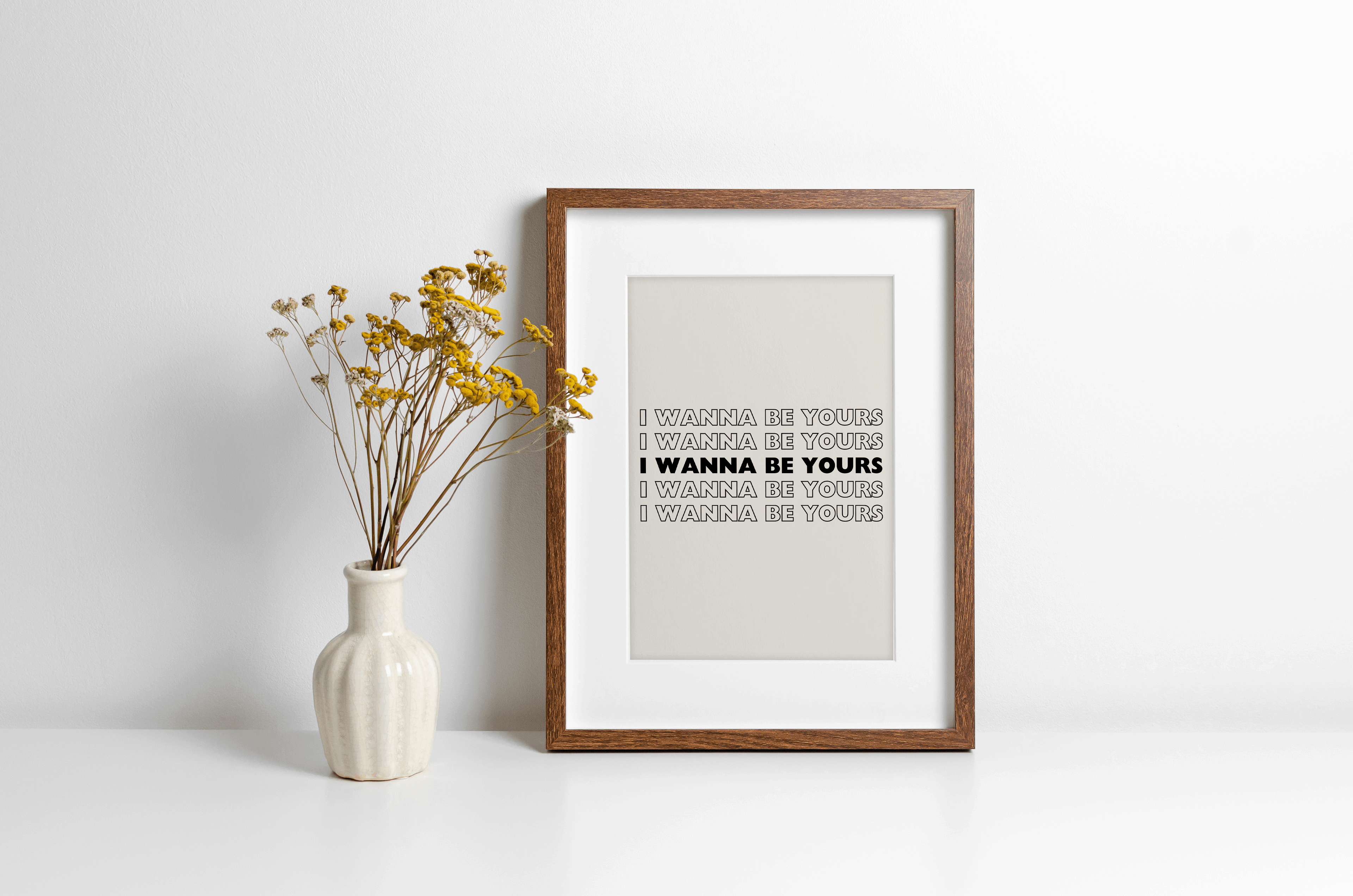 A music lyrics print featuring Arctic Monkeys "I wanna be yours" typography in black against a beige background. Displayed in a wooden frame against a white wall with yellow flowers in vase alongside.