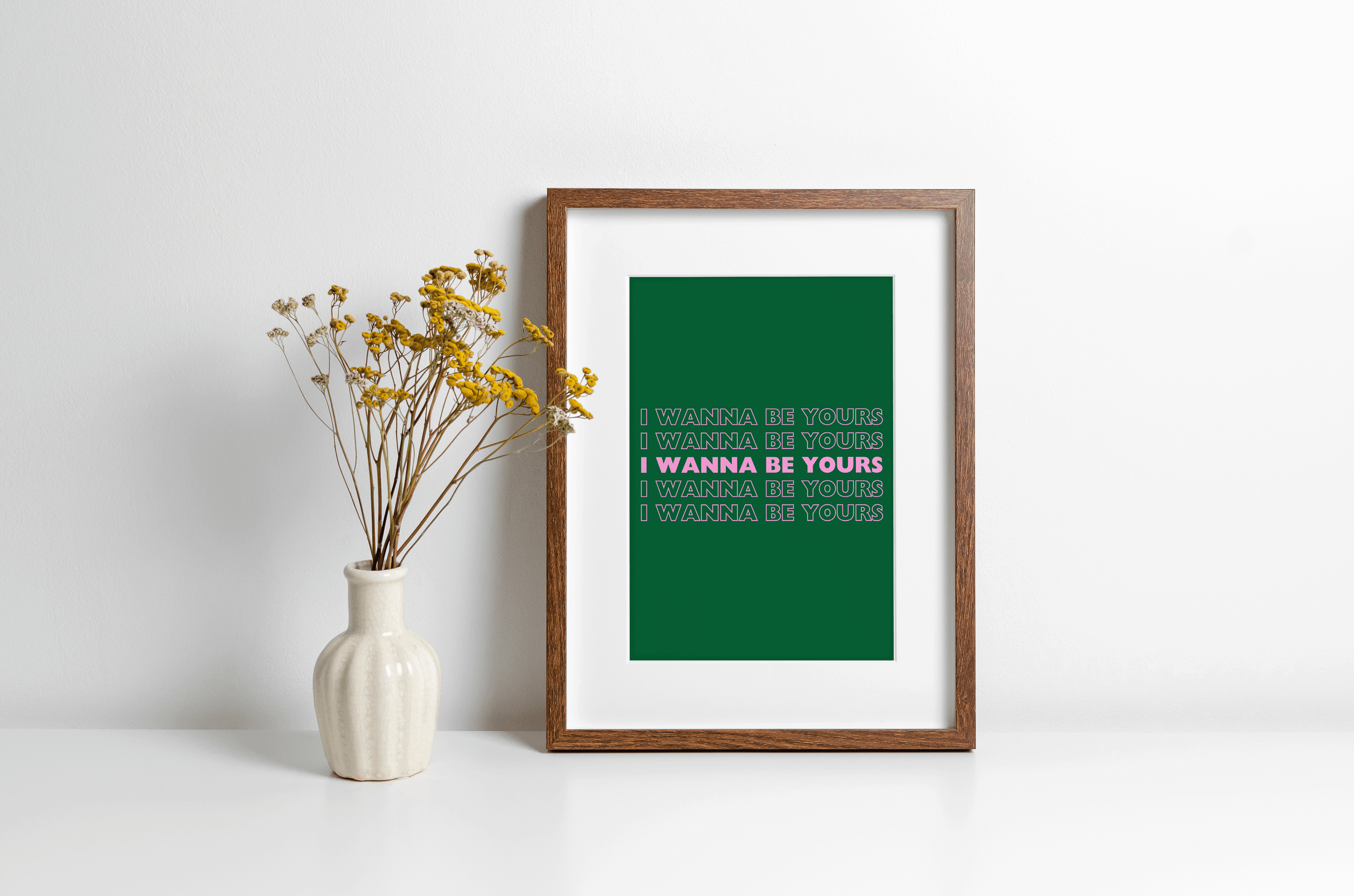 A music lyrics print featuring Arctic Monkeys "I wanna be yours" typography in pink against a green background. Displayed in a wooden frame against a white wall with yellow flowers in vase alongside.