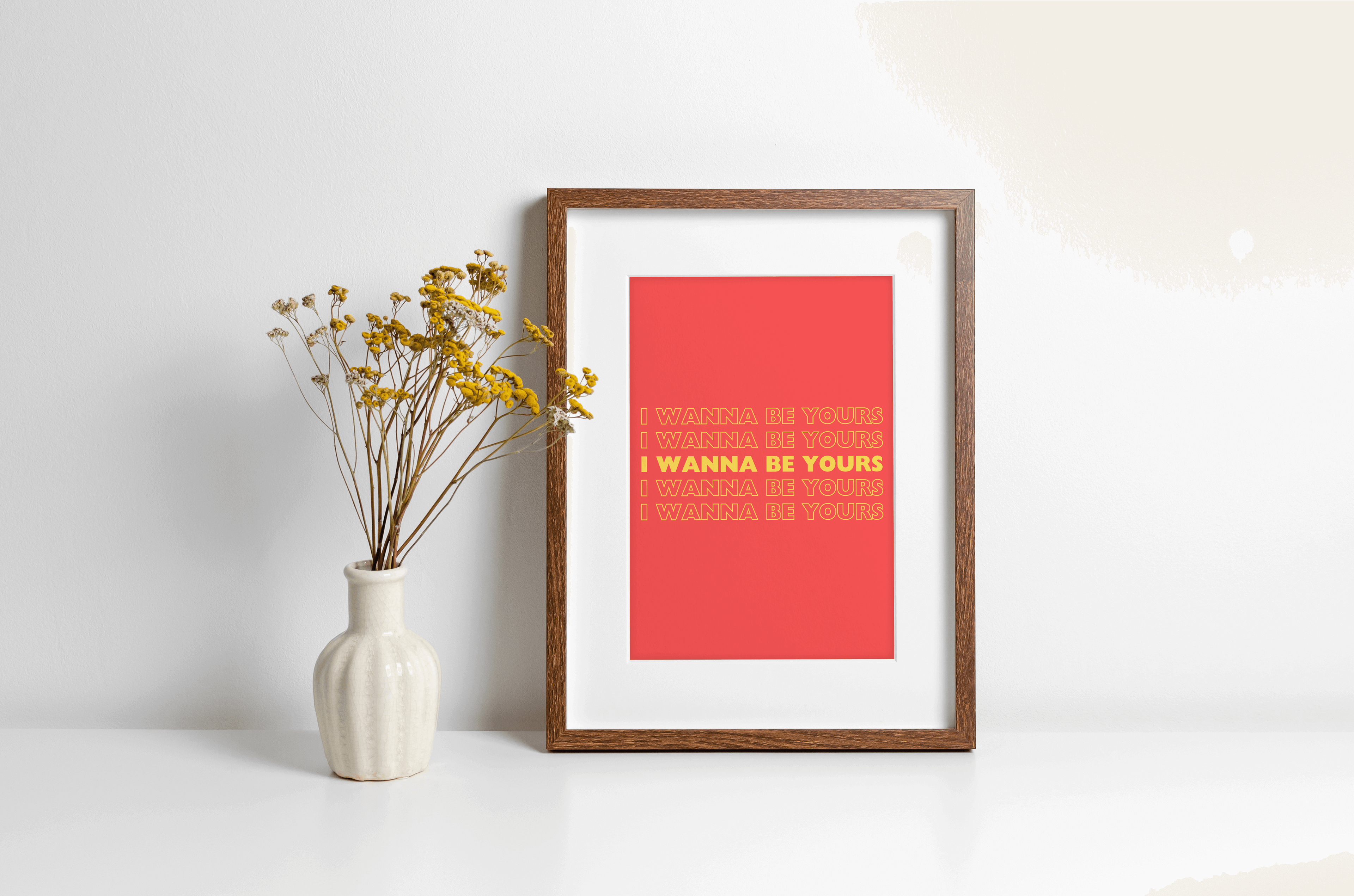 A music lyrics print featuring Arctic Monkeys "I wanna be yours" typography in yellow against a red-orange background. Displayed in a wooden frame against a white wall with yellow flowers in vase alongside.