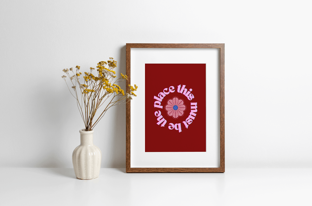 A bold red art print with pink text 'this must be the place' in a circular design around a flower illustration, displayed in a wooden frame alongside a vase filled with yellow flowers.