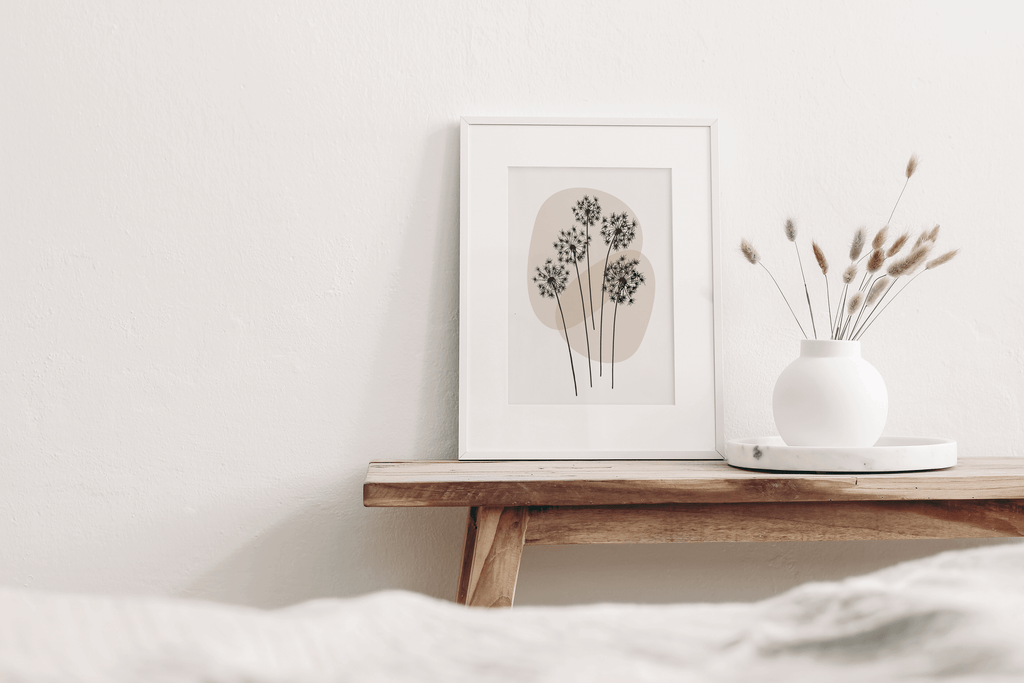 Minimalist line art print featuring a line drawing of dandelion, on a neutral background displayed in a bedroom. Print is shown in a white frame on a bench with decorative items.