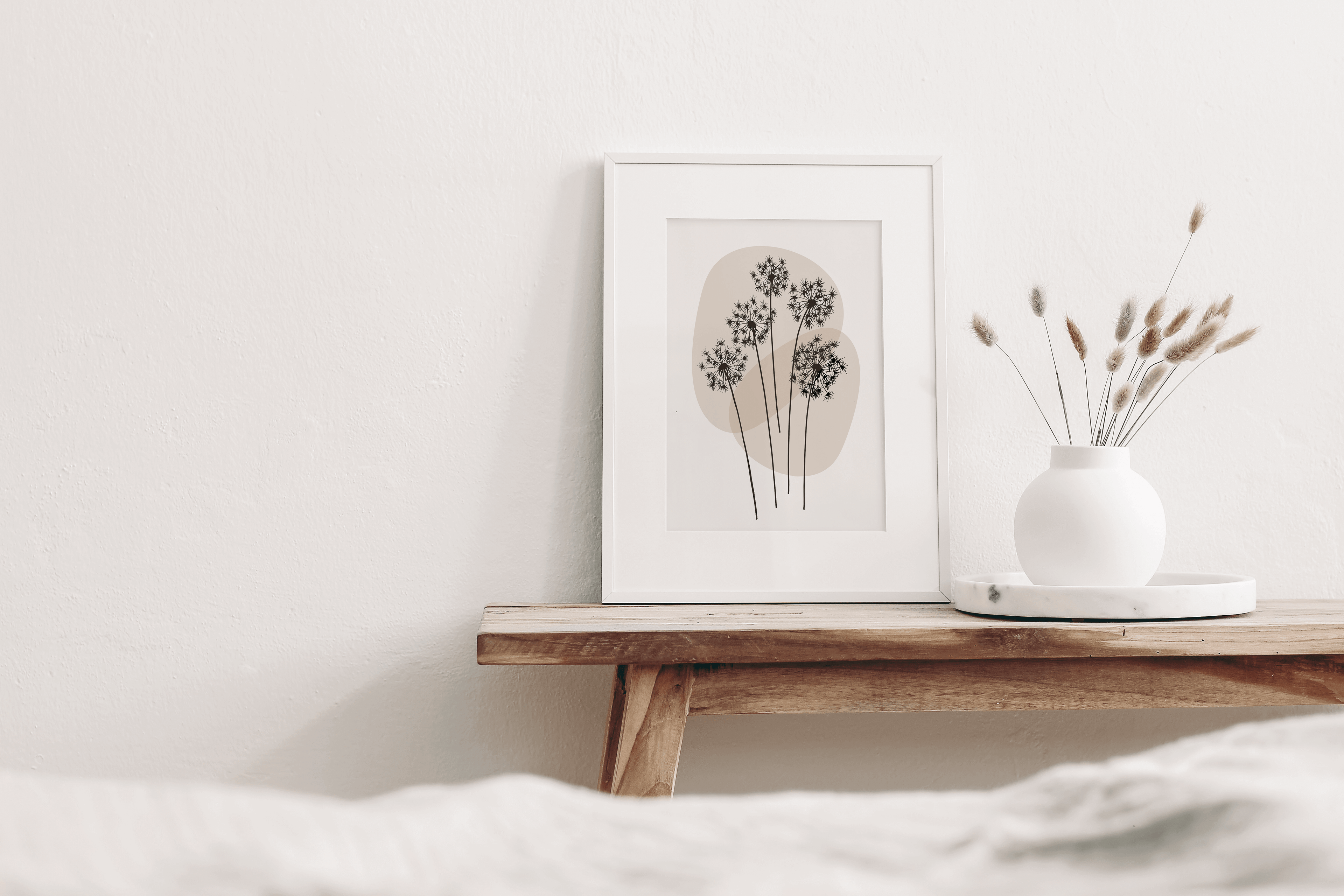 Minimalist line art print featuring a line drawing of dandelion, on a neutral background displayed in a bedroom. Print is shown in a white frame on a bench with decorative items.