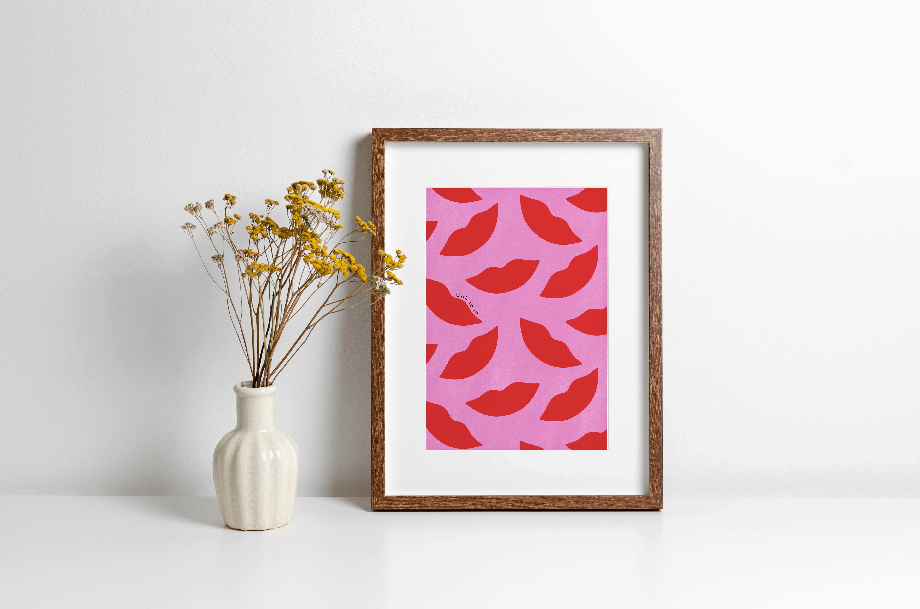 A bold art print featuring a red and pink lips pattern, displayed in a frame, with a vase of dried flowers to the side on a white surface.