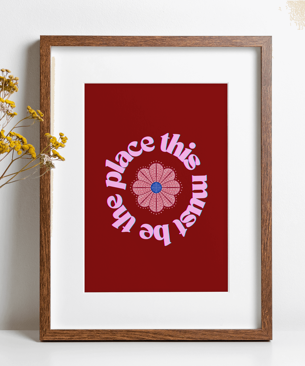 A red art print with pink text 'this must be the place' in a circular design around a flower illustration, displayed in a wooden frame.